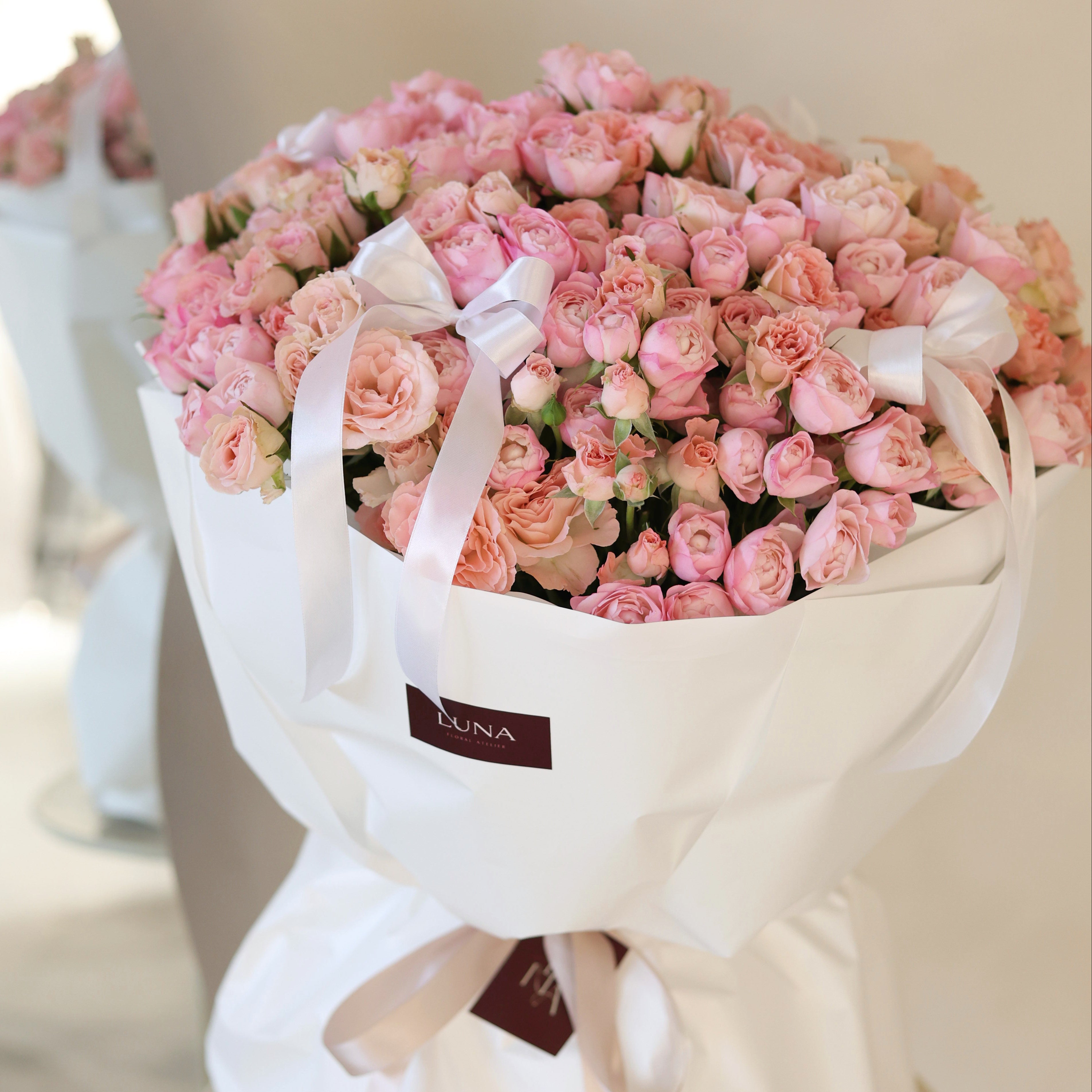 Bouquet of pink flowers wrapped in white paper with a visible brand label on a light background.